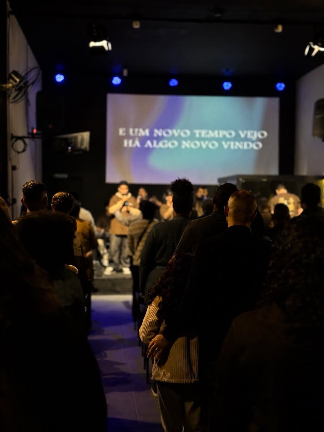 Congregação da Tetelestai Church em pé durante o louvor, com o telão e as luzes do palco ao fundo